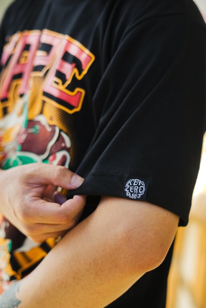 A man adjusts the sleeve of his black graphic T-shirt, showcasing a vibrant design and text logo.