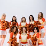 A group of eight smiling cheerleaders in vibrant uniforms holding shiny orange pom poms.