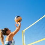 A woman plays volleyball on a sunny day, reaching to hit the ball over the net.